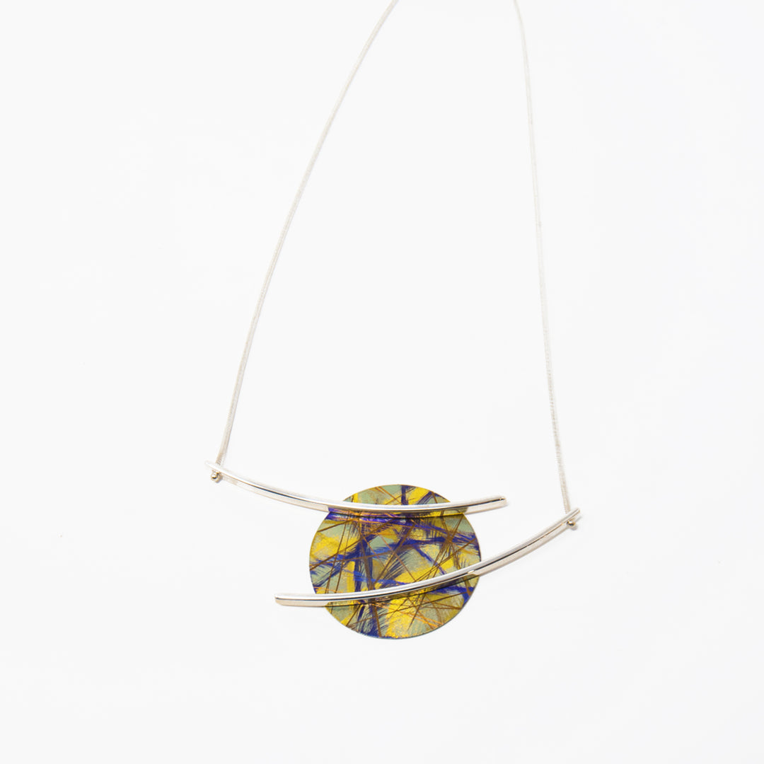 This titanium necklace is made from anodized titanium and sterling silver and has a circle shaped pendant. The circle is made from titanium and is yellow with colorful designs that are achieved by anodizing the metal. There are sterling silver branches stretching across and past the pendant, giving it a floating appearance. 