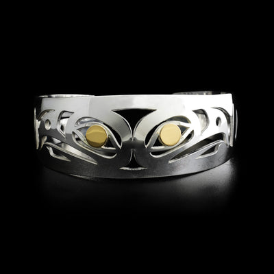 Side-view of two eagle heads, back-to-back. Laser-cut with spaces cut out. Both eagles have 14K gold in their visible eye. Minimalist design, by Tahltan artist Grant Pauls. Bracelet is sterling silver.