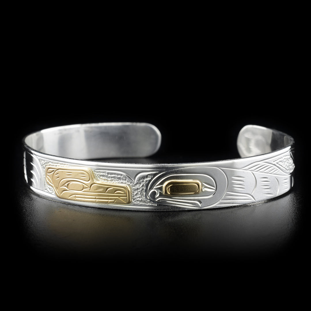This gold and silver bracelet depicts the head of a bear made from gold in the center facing a full-bodied salmon to its right that has a gold eye. The background has a stippling design.