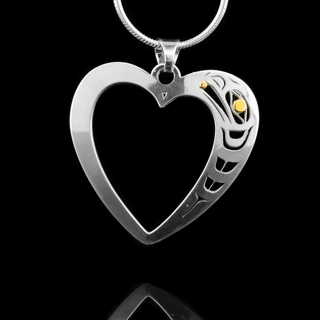 This heart pendant is made from sterling silver and 18K gold. The pendant is heart shaped and has a large heart shaped cutout in the middle of it. On the right side of the pendant, a depiction of the Raven has been cutout from its surface. The eye of the Raven and the pearl in its mouth have been made from 18K gold.