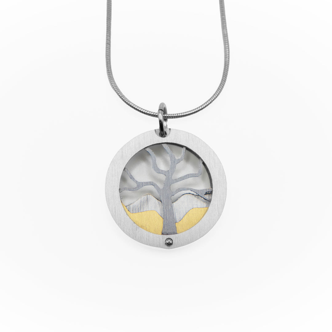 Silver-coloured frame with grey tree silhouette inside with silver-coloured and gold-coloured mountains in background. Brushed and anodized aluminum. By JR Franco. Stainless steel snake chain included.