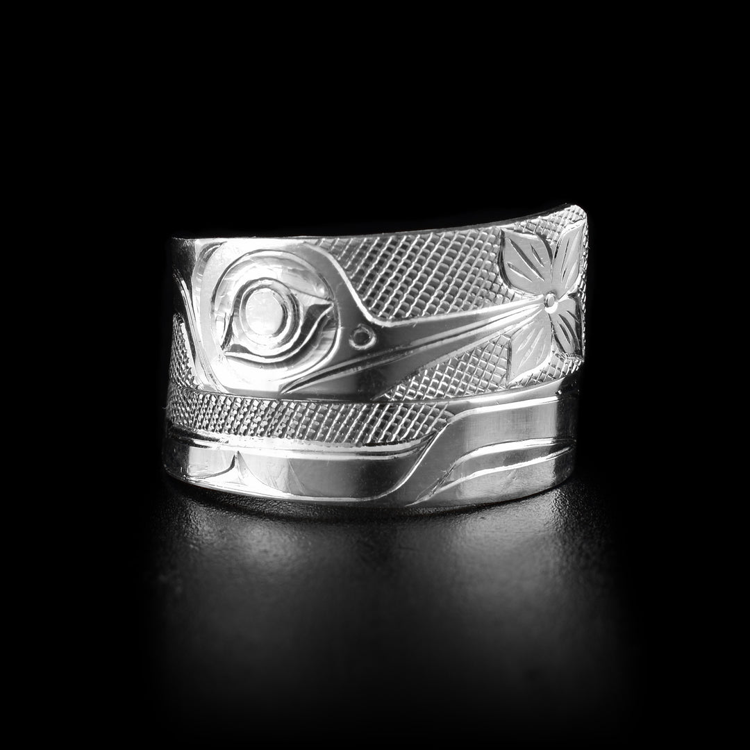 This sterling silver Hummingbird ring has a tapered band that wraps around the finger twice. There is a depiction of the side profile of the head of the hummingbird carved into the top, widest, band. The beak is facing the end of the band and there is a flower at the end of it. There are cross hatching designs in the background of the scene.