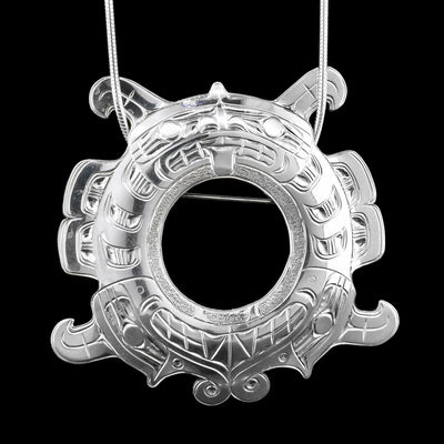 This Sisiutl pendant is made form sterling silver and has a large circular shape. The piece is shaped like a sea serpent the Sisiutl and has two heads reaching around to for a circle. There is a large circle cutout in the middle of the piece and carvings on the surface of is depicting the Sisitul.