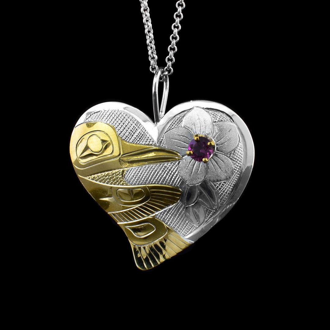 This heart-shaped Silver and Gold Hummingbird Pendant is ane with sterling silver and the hummingbird legend plated in gold. It faces towards a flower and inside the hand carved flower is a violet garnet.