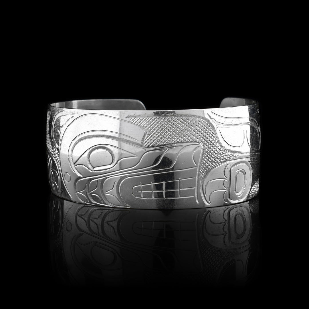 This Wolf bracelet has a wide, sterling silver band with a gap in the back. The head of the Wolf is carved in the center of the piece in a side profile and takes up the width of the band. To the right, there is one of its clawed paws and its tail curling up to the top of the band. On the left its other leg is shown.