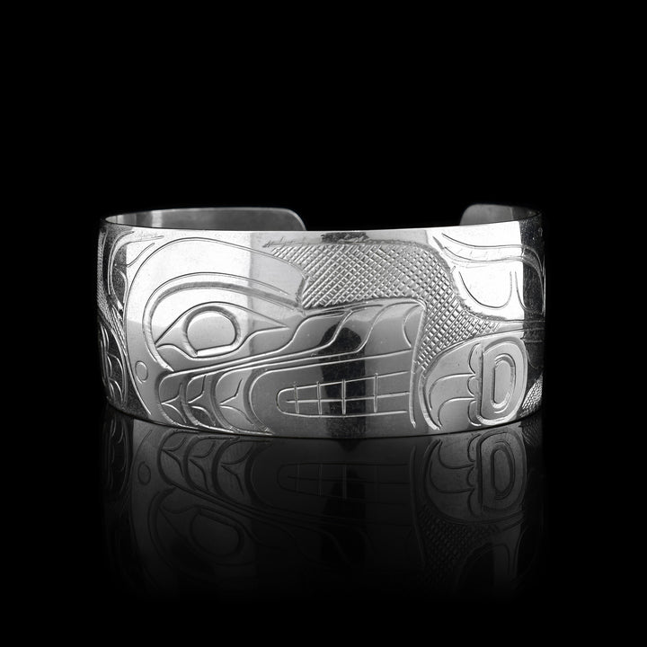 This Wolf bracelet has a wide, sterling silver band with a gap in the back. The head of the Wolf is carved in the center of the piece in a side profile and takes up the width of the band. To the right, there is one of its clawed paws and its tail curling up to the top of the band. On the left its other leg is shown.
