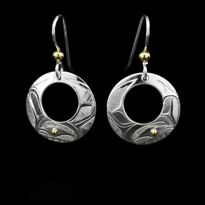 These sterling silver earrings have circle hangs with circle cutouts. There are depictions of the Orca carved into the surface. There are 14K gold balls in the eyes of the Orca and attached to the hooks.