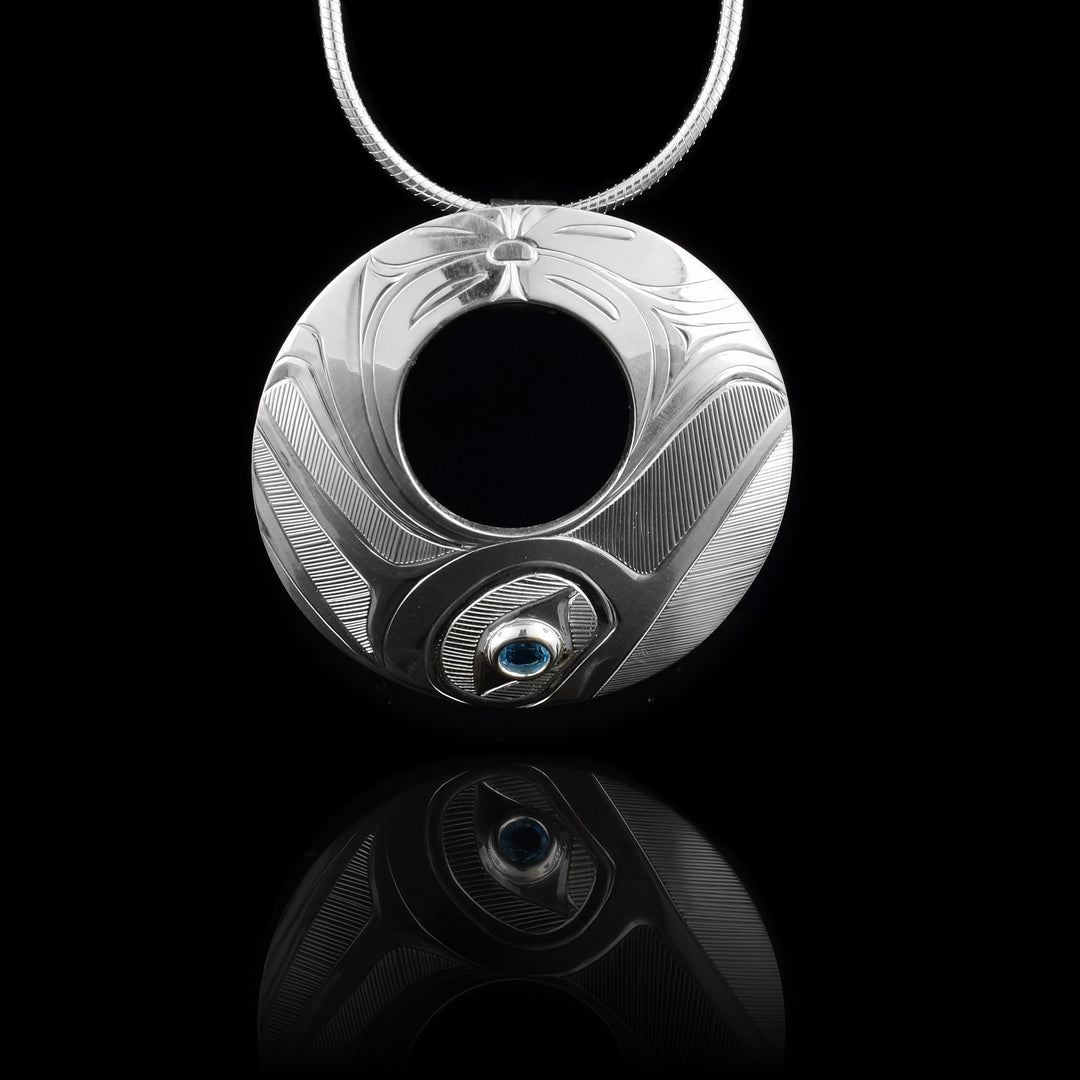 This pendant blue topaz is made from sterling silver and is circle shaped with a small, circle cutout in the center of the piece. There is a depiction of the Hummingbird carved into the bottom of the piece and a flower design made in the top of it. The blue topaz is held at the bottom in the eye of the Hummingbird. Its beak extends to the right and its wing on the other side.