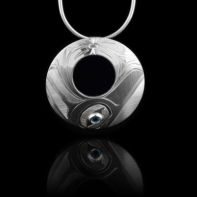 This pendant blue topaz is made from sterling silver and is circle shaped with a small, circle cutout in the center of the piece. There is a depiction of the Hummingbird carved into the bottom of the piece and a flower design made in the top of it. The blue topaz is held at the bottom in the eye of the Hummingbird. Its beak extends to the right and its wing on the other side.