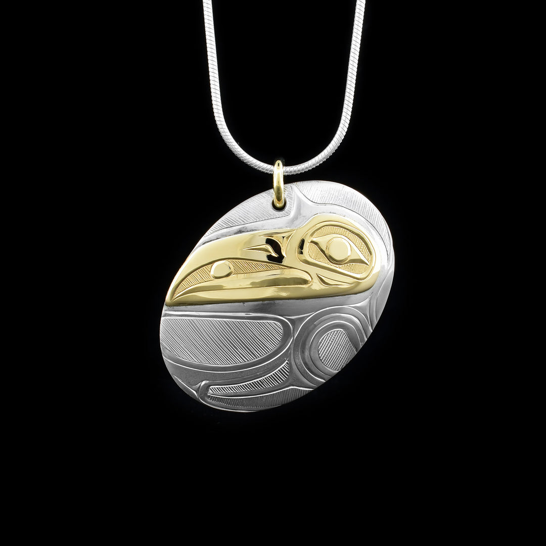 This Raven pendant is made from sterling silver and 14K gold and has an oval shape. The pendant is chunky and has carvings of the surface of it that depict the Raven and the head of the Raven is made from gold.