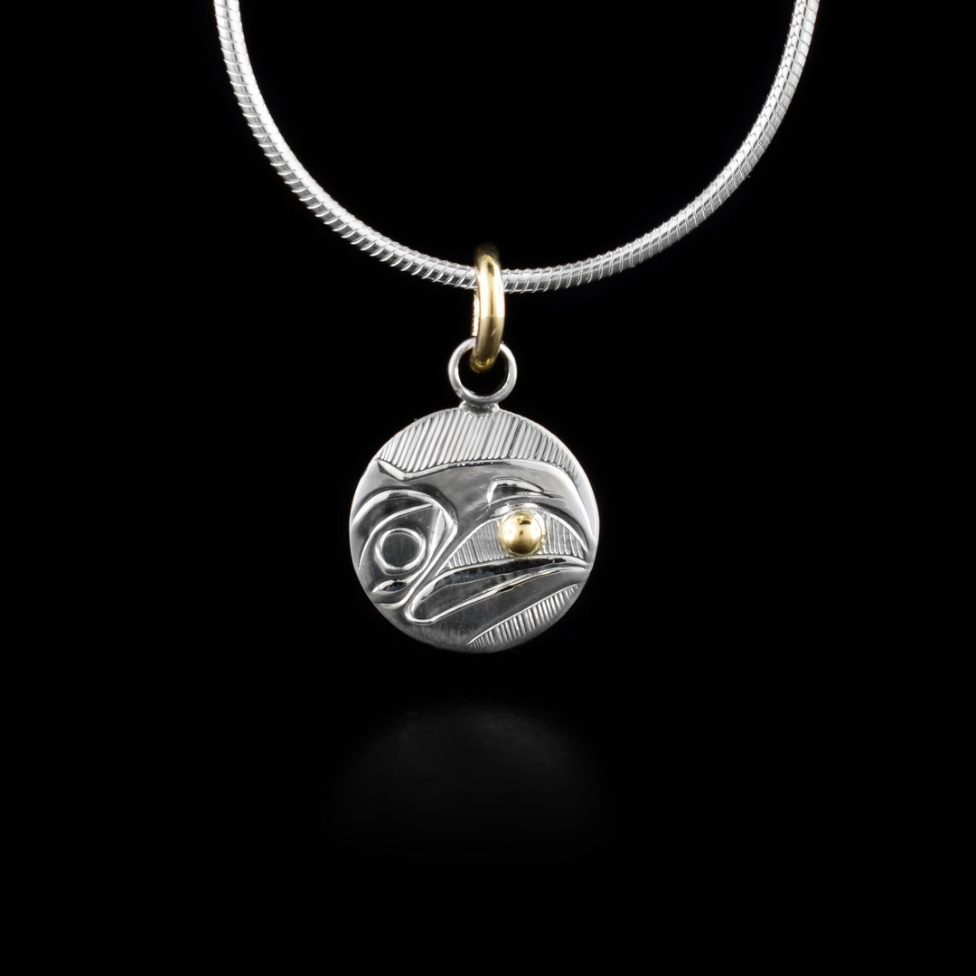 This Raven pendant is made from sterling silver and has a small, circle shape. The head of the Raven is depicted in a side view in the center of the piece. Its beak is open and there is a 14K gold stud held between it. Behind the head there are hatching patterns.