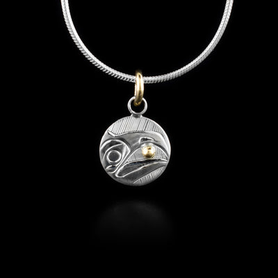 This Raven pendant is made from sterling silver and has a small, circle shape. The head of the Raven is depicted in a side view in the center of the piece. Its beak is open and there is a 14K gold stud held between it. Behind the head there are hatching patterns.