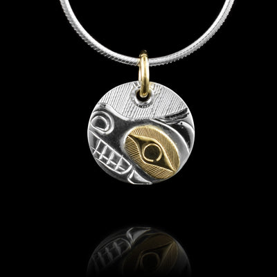 This 14K gold Bear pendant is small, circular and made from sterling silver. The piece hangs from a 14K gold jump ring and there is a carving depicting the bear legend on the surface of it. The side view of the Bears head is shown and a portion around its eye is done using gold There are hatching patterns behind the head.