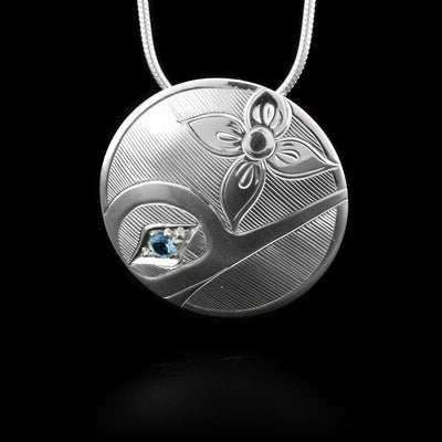 This blue topaz pendant is circle shaped, made from sterling silver, and has carvings that depict the Hummingbird on it. There is a blue topaz gemstone embedded in the eye of the Hummingbird. 