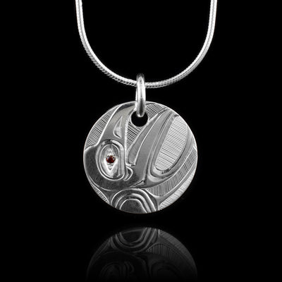 This Sterling Silver Round Raven pendant with Garnet is hand carved by artist, Hollie Bartlett. The Raven is carved into the piece at a side profile. Its head and a piece of its wing are shown and there are fine, symmetrical lines in the negative space of the scene.