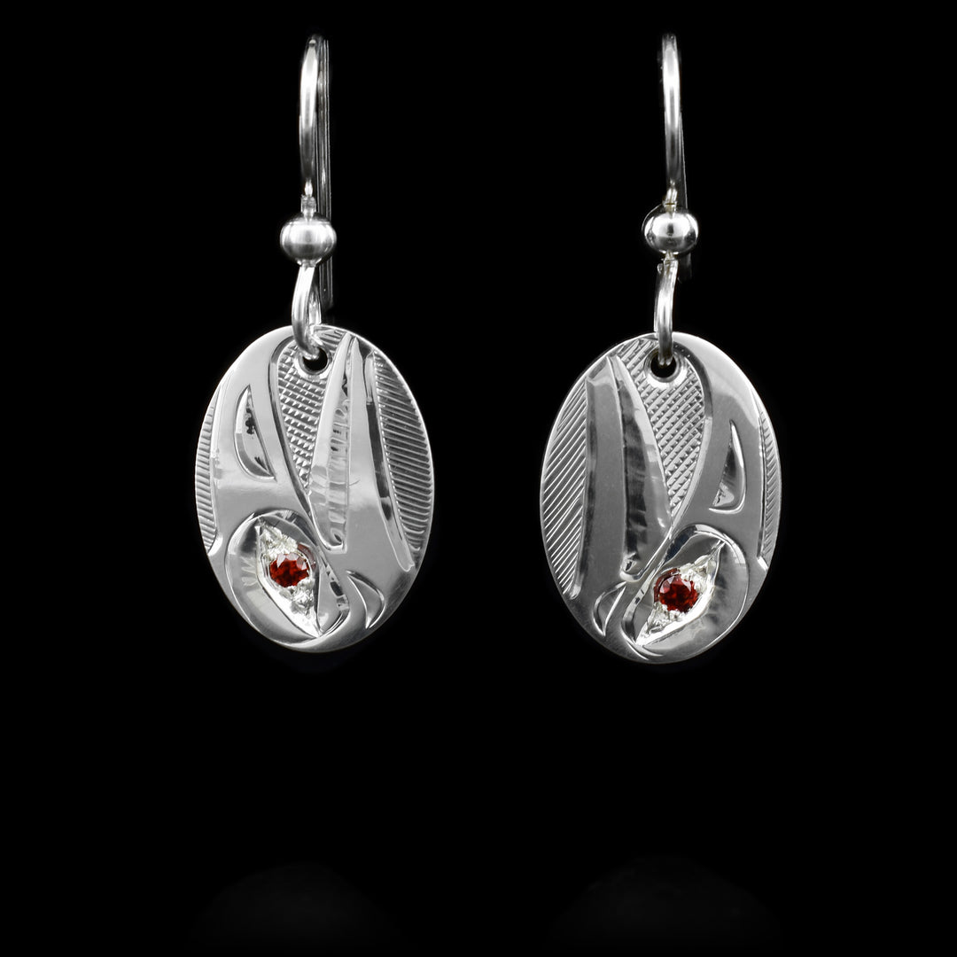 These oval, silver dangle earrings are made from sterling silver and has a depiction of the Raven carved into it. There are garnet gemstone embedded in the eyes of the Raven.