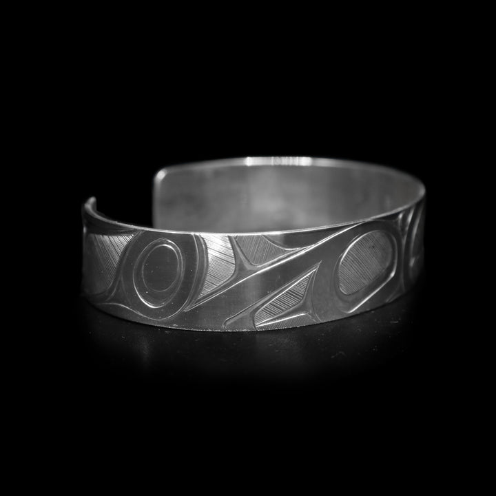 This Orca whale bracelet has a single, sterling silver band with a gap in the back. There is a depiction of the Orca legend intricately carved into the surface of the band. The head of the legend is in the center of the band and its fins are depicted on either side of the head. The accents are made with fine, hatching patterns.