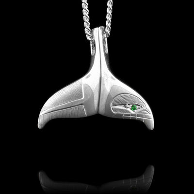 This sterling silver whale tail necklace pendant is shaped like a whale's tail. On the right fin, there is a simple depiction of the head of the Orca with its eye and teeth. On the other fin, there are fine, symmetrical lines representing its tail. There is a green tsavorite gemstone set into the eye of the Orca.