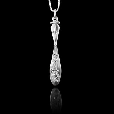 This Raven pendant is made from sterling silver and has a thin, long shape. The piece is shaped like a paddle from a boat and there is a depiction of the Raven legend carved into the pendant. The head of the Raven is on the oar portion and is facing upwards.