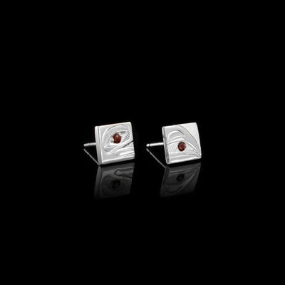These square stud earrings are made form sterling silver and there is a depiction of the Raven's head carved across the two earrings. There is a garnet gem in one depicting the eye of the legend and in the other that is held between the beak of the legend.