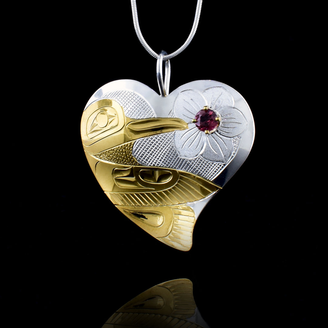 A hand-carved sterling silver heart featuring a vibrant hummingbird in 18K gold-plating. A deep red garnet gemstone is set at the center of a carved flower, drawing the eye to the intricate textures of the piece.