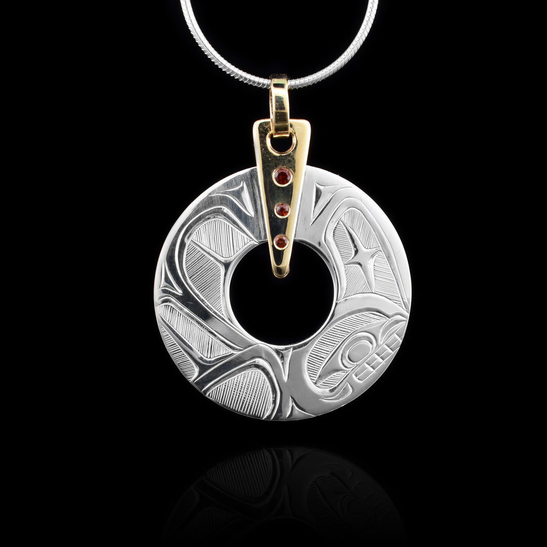 Hummingbird & Orca Pendant is crafted from sterling silver; this circular pendant features carved designs of an orca and a hummingbird. The attachment is a 14k gold triangle set with three red garnets.