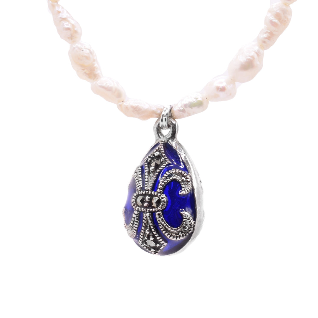 This pearl bead necklace has pearls that get smaller towards the back of the piece. There are small rows of blue highlight beads at the very back of the necklace. There is a small oval pendant made from blue crystal that has silver charm designs over top of the crystal. The charm has a silver band around the circumference and flowers designs on it.