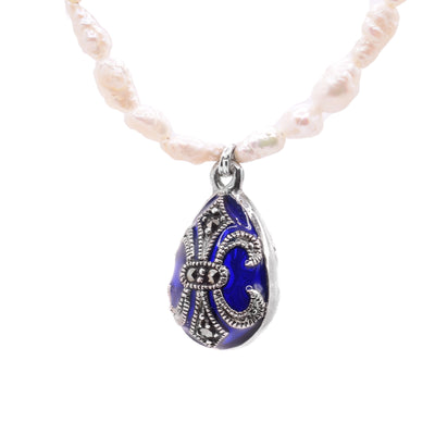 This pearl bead necklace has pearls that get smaller towards the back of the piece. There are small rows of blue highlight beads at the very back of the necklace. There is a small oval pendant made from blue crystal that has silver charm designs over top of the crystal. The charm has a silver band around the circumference and flowers designs on it.