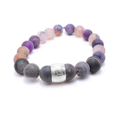 This beaded purple agate bracelet is made up of multi colored agate beads. The beads are sphere shaped, small and have tan, purple and dark colors on them. There is a sterling silver spirit bead in the front with the darker beads around it. There is a depiction of the head of the Orca carved into the spirit bead.