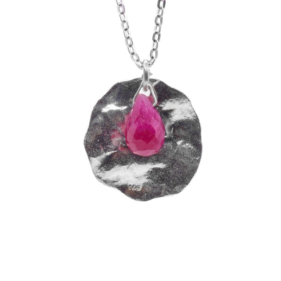 This ruby pendant necklace has a circle shaped, sterling silver pendant that has a crumple look to it. The edges of the circle are uneven and rough in align with the crumpled appearance. In front of the piece there is a teardrop shaped ruby gemstone dangling from a jump ring.