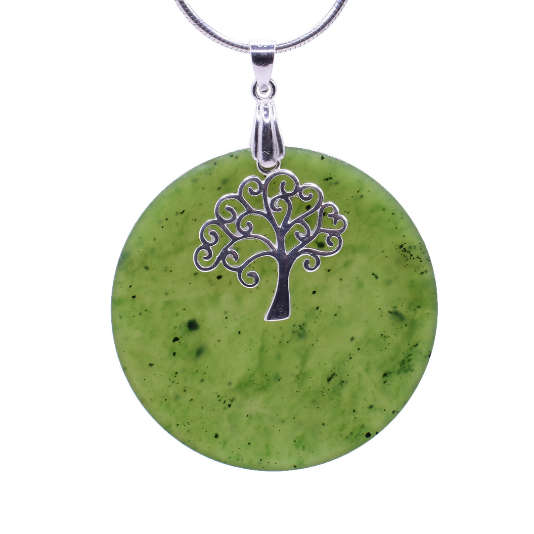 This jade disc pendant has a flat, circle shaped jade piece with a small, silver tree charm resting in front of it. The piece hangs from a silver bail. The jade is a light green and the branches of the tree somewhat resemble heart shapes.
