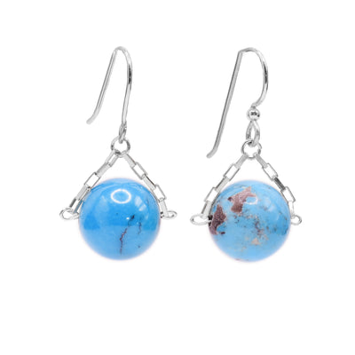 These turquoise drop earrings have small, sterling silver chains attached to the hooks and to either side of the turquoise hangs. The turquoise are large spheres and dangle from the small chains.