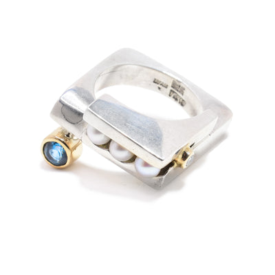 This sterling silver ring has a square shape. Attached into the signet are three freshwater pearls. Attached to the ring with 14K gold is a blue topaz gemstone and a diamond on either end of the ring.