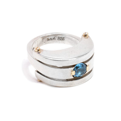 A sterling silver band with a chunky front. There is a round blue topaz stone in the middle. There are two gold details on the sides of the stone, and four in the top and the bottom of the chunky part.