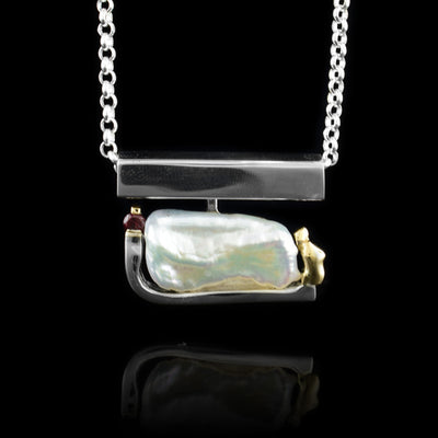 This baroque pearl pendant has a large, pearl that is held, horizontally with a sterling silver arm. The pearl lays below a silver bail that is a long and there are 14K gold details on either end of the pearl. On one side of the piece is a small, garnet gemstone.