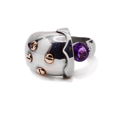 This sterling silver ring has an abstract shape with one end of the band being very large and round with designs on it. There are 14K gold studs on the large part of the ring and an amethyst gem held between the band.