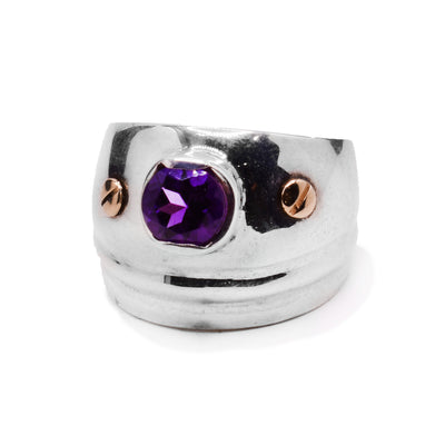 This sterling silver amethyst ring has a wide band and is tapered. At the top of the ring is an embedded amethyst gem with a 14K gold stud on both sides.