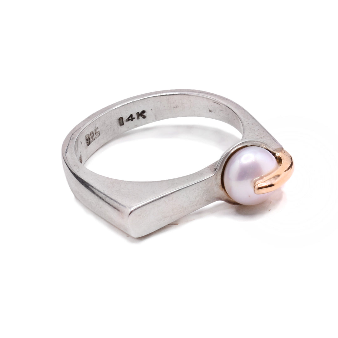 A sterling silver pearl ring with a flat center. On the flat side, there is a white freshwater pearl that sits off center. There is a 14K yellow gold detail hugging the pearl.