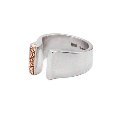 A sterling silver band that is tapered and split in the middle. One of the  sides of the band that is split has a vertical 14K gold detail on it that has a crosshatching pattern.