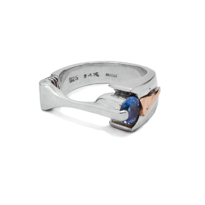 This sterling silver ring has a thin, flat part of the band in the front. There is a blue topaz gem on the side. There is a yellow gold part on the band going around the finger.
