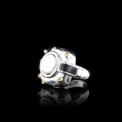 This chunky pearl signet ring has a wide, sterling silver band and at the top of the ring there is a large, rounded square made from silver. There is a large, freshwater pearl in the center of that square. There are 14K gold studs around the pearl with a blue topaz and garnet gem there too. Extending from the pear, there are rectangle, plexi glass pieces that contrast the pearl.