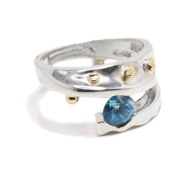 A sterling silver band with a silver branch that coils halfway around the band. Between the band and the silver branch, there is a round blue topaz stone. There are four 14K gold bolt details along the band.