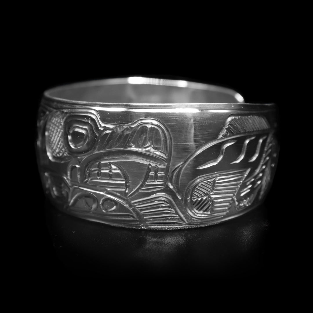 This sterling silver cuff bracelet has a wide band with a gap in the back. There are depictions of multiple indigenous legends carved into the surface of the band.