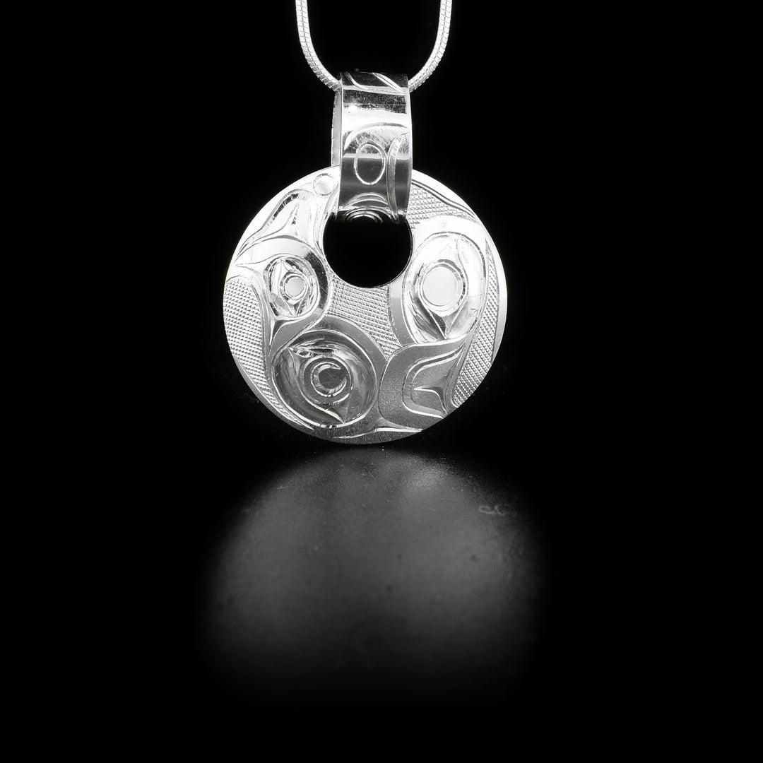 This sterling silver pendant is circle shaped and includes a small, circle cut out and a hand carved bail running through the cutout. There is a double Eagle pattern carved into the pendant.