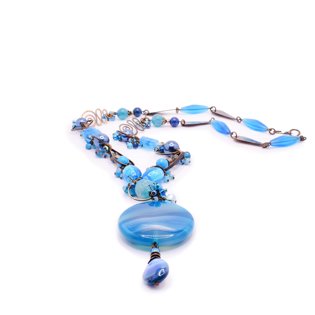 Artina's has this Honica statement necklace featuring a stunning blue-hue design. It showcases a Mother of Pearl drop adorned with Apatite and Dyed Agate, accented by Austrian Crystals and Lampwork glass on an antique brass chain.