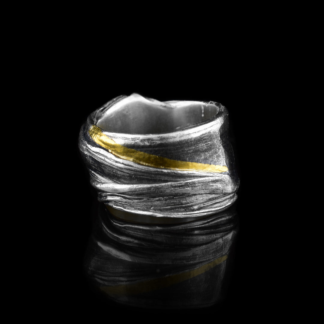 This wavy ring band is made from sterling silver and has flowing designs on the surface of it. The piece is made by casting metal into wax so it mimics the appearance of its wax mold. There are keum boo gold highlights on the band around one of the strips of sterling silver and the band is wide.