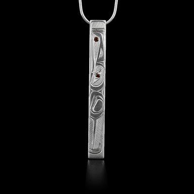 Long, thin rectangular sterling silver pendant with side-view of raven. A garnet has been set in the eye and in between the beak. Hidden bail on back. Hand-carved by Haisla artist Hollie Bartlett.