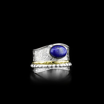 This spinner ring is made form sterling silver, brass and lapis lazuli gemstone. There is a wide, wavy and silver band with an ovoid lapis gem held on the top of it, There is a textured silver hoop and a brass hoop on the band that spin around the band.