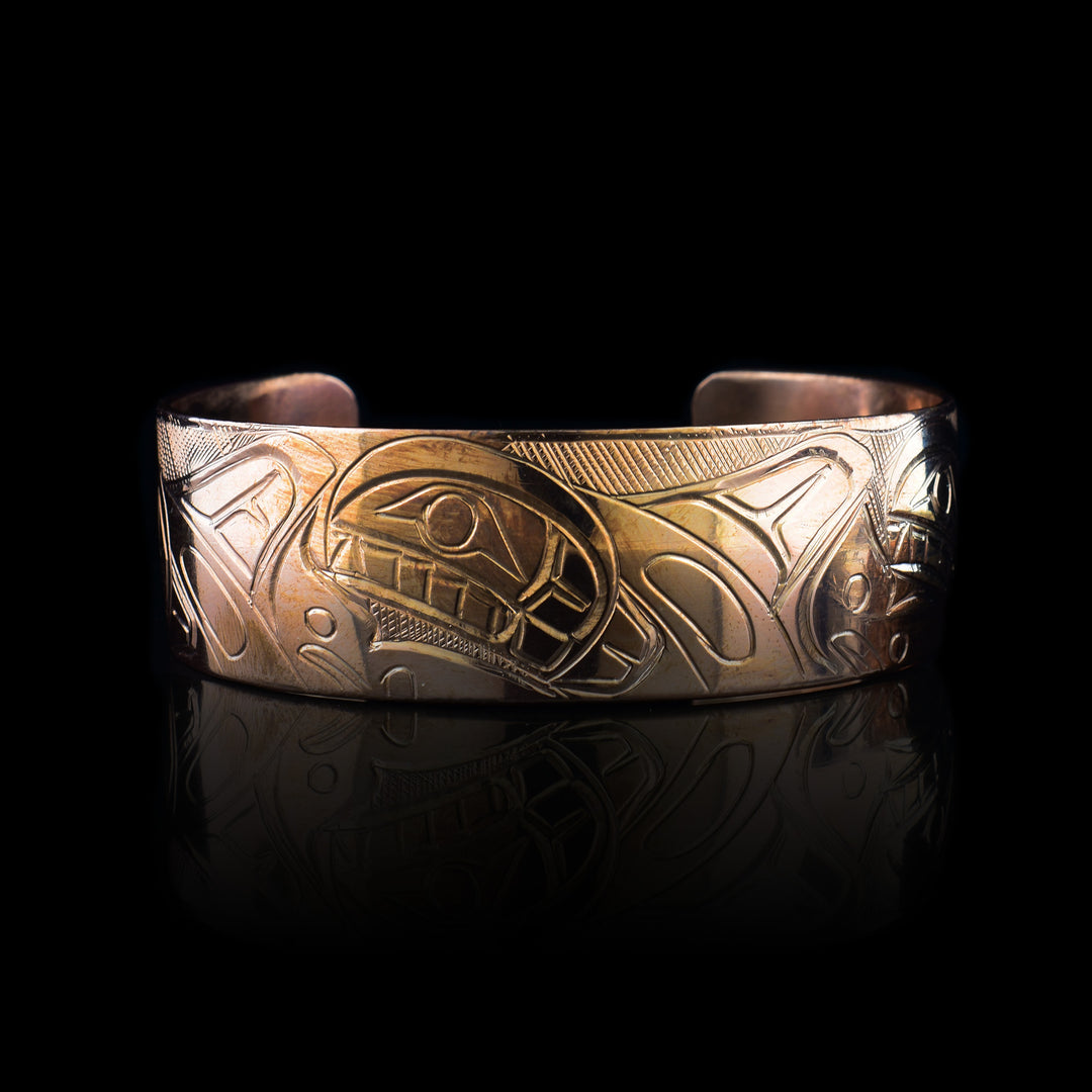 This copper cuff bracelet has a wide band with a depiction of three, Orca whales carved into the surface of it. The whales are done at a side view and are breaching with their heads and Doral fins shown. There are splashing designs of the ocean made around the Orca pod.