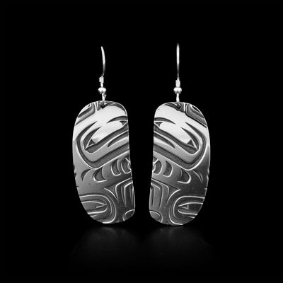 Laser-carved ovoid-shaped dangle earrings. Oxidized background emphasizes three-dimensional design. Abstract design in both earrings depicts an orca. By Tahltan artist Grant Pauls.